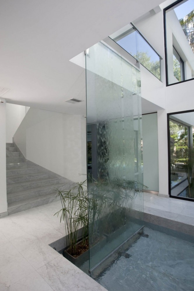 To continue the inside-outside feel, the architect has included a decorative indoor pool as part of a large yet understated water feature which reflects an adjacent pool out on the patio; a waterfall against a transparent pane of glass cascades all the way from the second floor, where the ceiling is cut away to reveal the upper living space, and a row of plants breathe greenery into the home, reflecting the garden beyond.