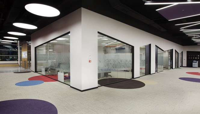 Beneath a chaotic and colorful designed ceiling, a circle carpet pattern decorates the floor, which is a familiar sight from other branch offices where it is used to reflect the company's corporate identity.