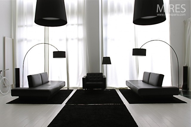 A symmetrical furniture layout creates a mirrored effect in the lounge area, complete with matching minimalistic sofas and slick black arc lamps to illuminate each side equally. This symphony of silhouettes stretches out across the white floor where a base of black throw rugs are perfectly aligned like a bridge over to, and beneath, the seating, and continues all the way up to the ceiling in the matching ebony pendant light shades.