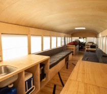 This photo shows the entire length of the converted bus home. The furniture is simple wood and the floor is made of reclaimed gym flooring. None of the structures built go above the window line keeping it linear and open to outside light.
